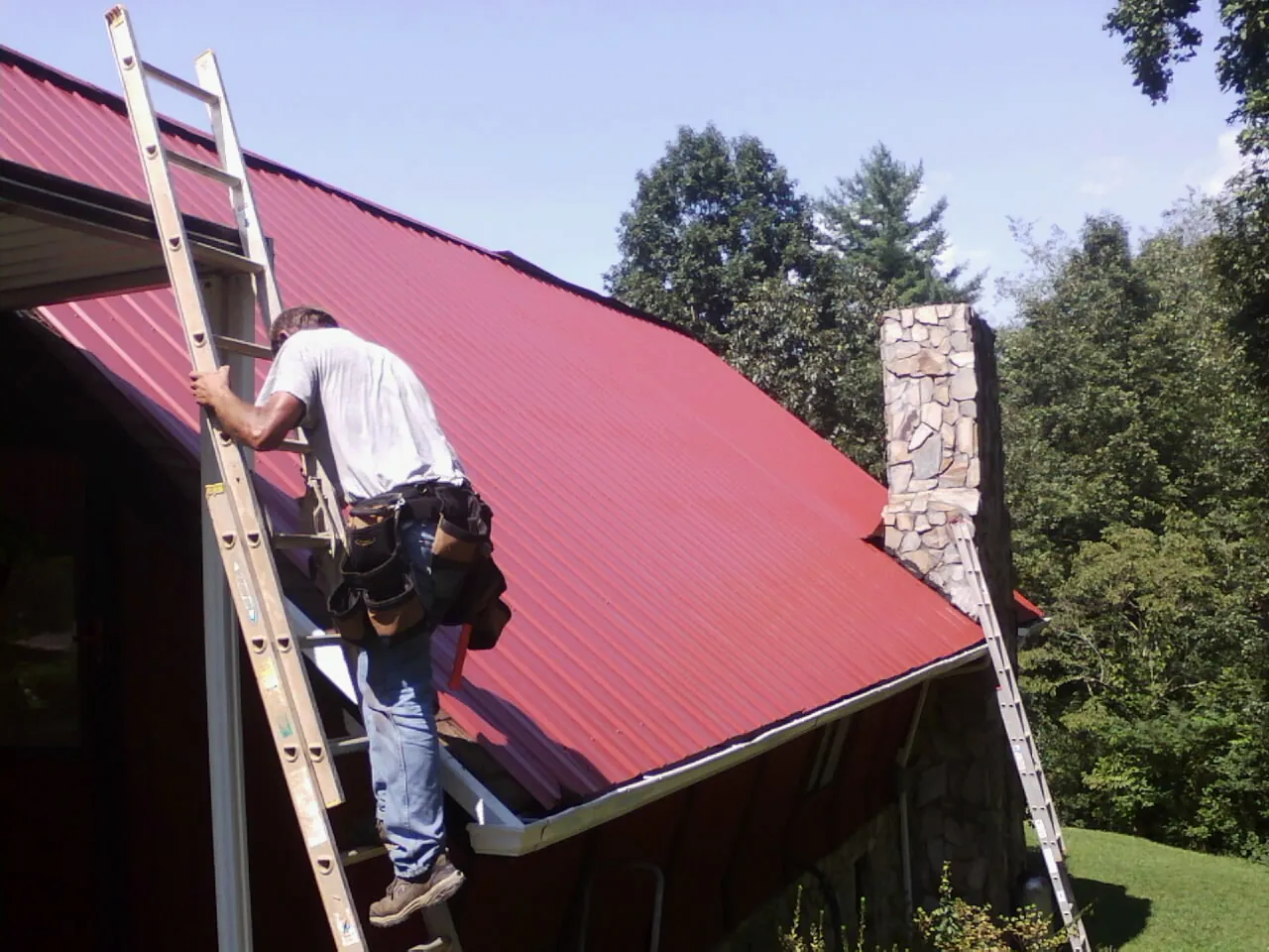 Roofer on a ladder beside a red metal roof and stone chimney — Soffit & Fascia Repair by Roofing Brothers in Shrub Oak