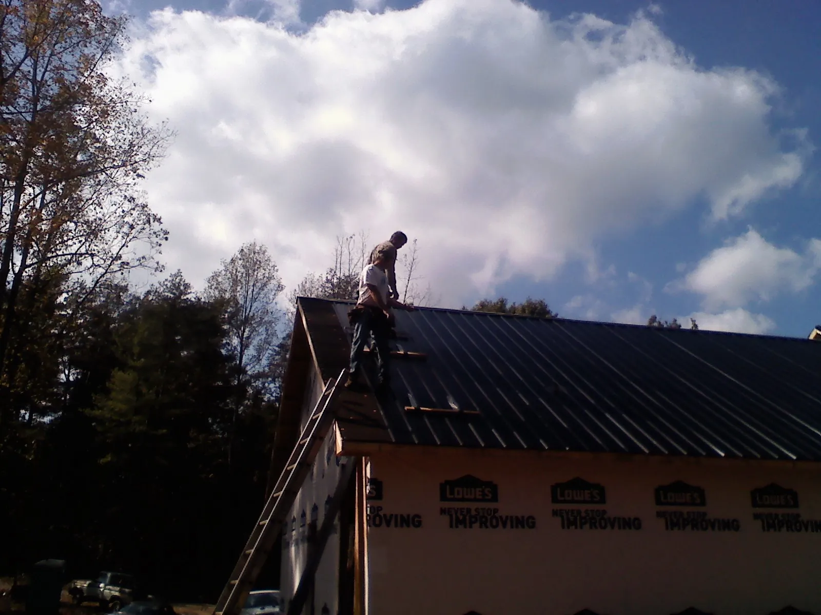 Roofer fastening dark metal panels high against a cloudy sky, from a Roof Inspection job in Shrub Oak